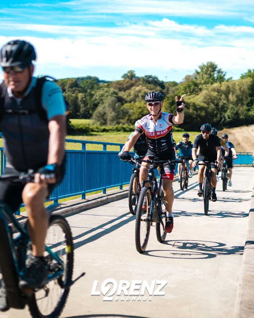 Vier Radfahrer in Trikots und Helmen auf einer sonnigen Brücke; der mittlere winkt in die Kamera.