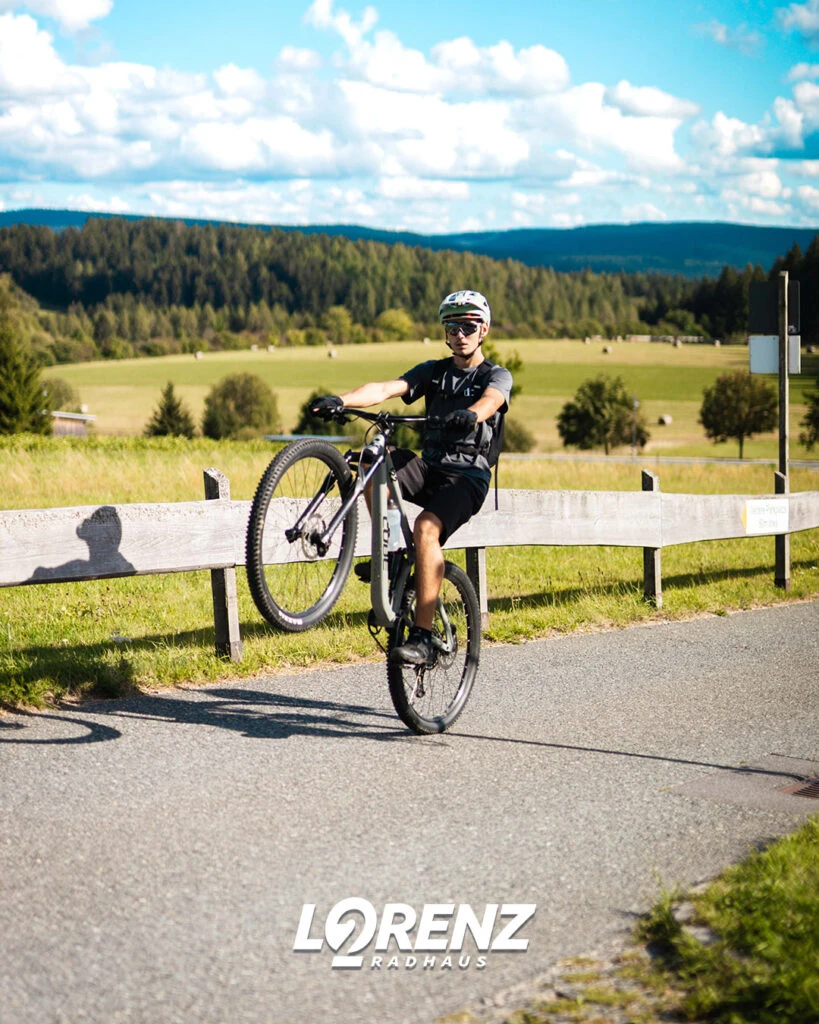 Ein Radfahrer macht auf einer asphaltierten Straße einen Wheelie vor einer ländlichen Kulisse.