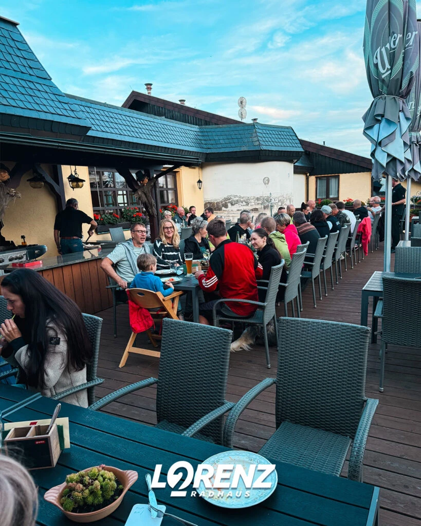 Eine große Gruppe von Gästen sitzt auf einer Restaurantterrasse an langen Tischen im Freien.