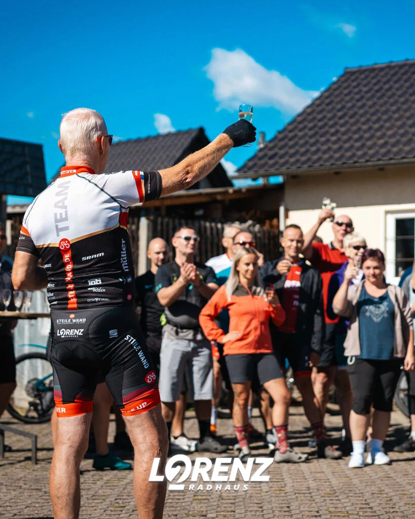 Ein Radfahrer reckt mit einem Sektglas in der Hand den Arm hoch, während eine Gruppe applaudiert.