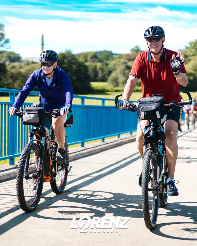 Zwei Radfahrer auf einer blauen Brücke, die während der Fahrt in die Kamera winken.