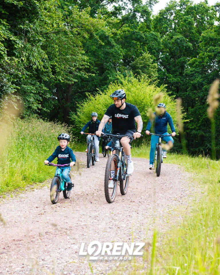 zwei Väter und zwei Kinder fahren auf E-Bikes und auf Kinderrädern durch die natur